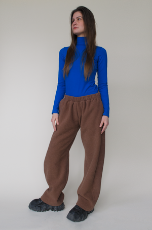 woman standing against beige backdrop wearing a cobalt blue turtleneck and brown straight-leg, low-rise sweatpants. paired with black sneakers. Long brown straight hair and fair skin. 