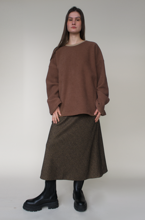 woman standing in front of grey backdrop wearing a large oversized brown fleece sweater, and a long brown wool skirt with black boots. Light fair skin with long brown hair.