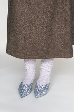 A pair of women's feet in cream-colored fuzzy socks, paired with shiny silver high heels and a long flowing brown skirt.