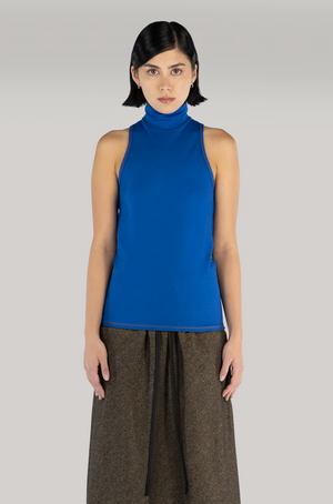 A woman standing in front of a grey backdrop wearing a cobalt blue sleeveless turtleneck top paired with a brown wool maxi skirt. model is light skin with short black hair.