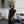 Load image into Gallery viewer, young woman standing in fashion studio wearing a Black one-shoulder tank top, with a band that crosses over the chest and arm. Design by Alex Watson Studio.
