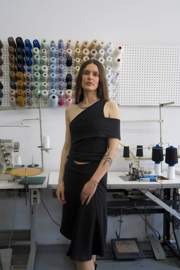 young woman standing in fashion studio wearing a Black one-shoulder tank top, with a band that crosses over the chest and arm. Design by Alex Watson Studio.